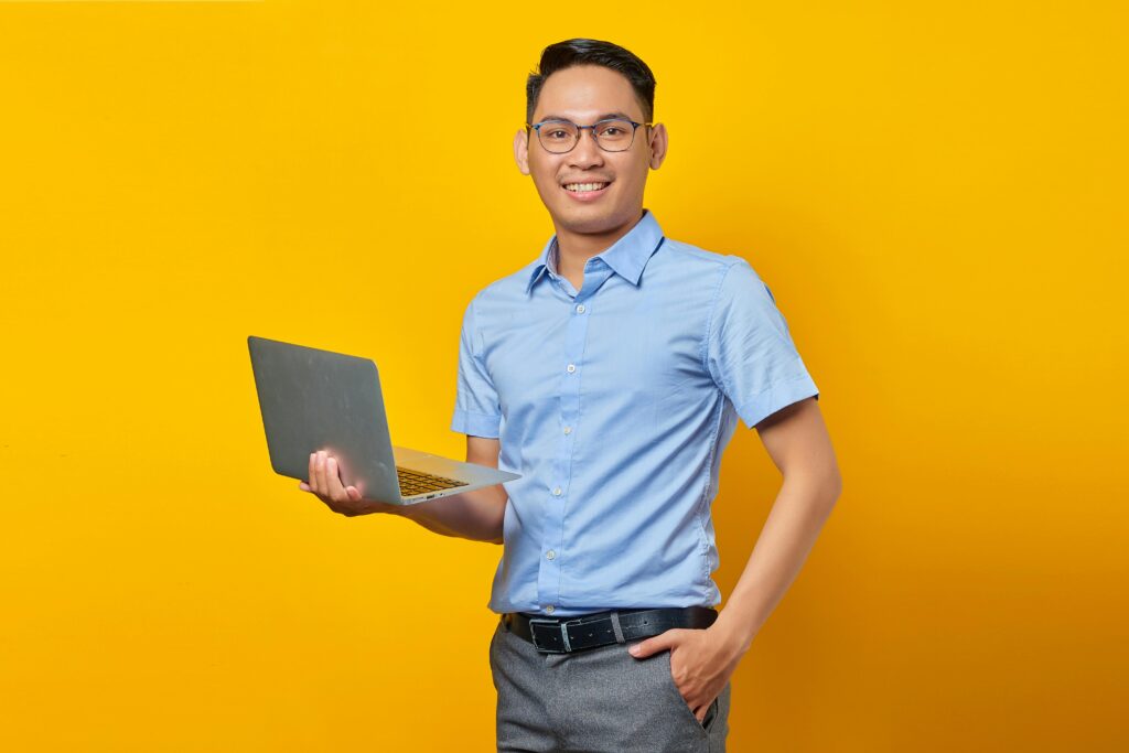 A confident young adult male with eyeglasses, holding a laptop against a vibrant yellow background, exuding a professional vibe.