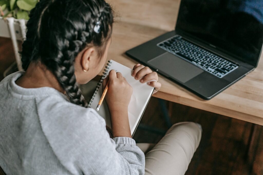 Young girl writing in a notebook with a laptop, showcasing focused learning.